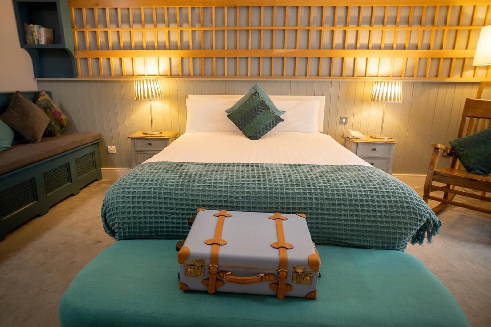 A neatly arranged bedroom with a bed, bedside lamps, a small table, a chair, and a suitcase on an ottoman.