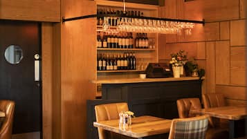 A restaurant interior with wooden tables and chairs, a bar area with wine bottles and glasses, and hanging wicker chandeliers.