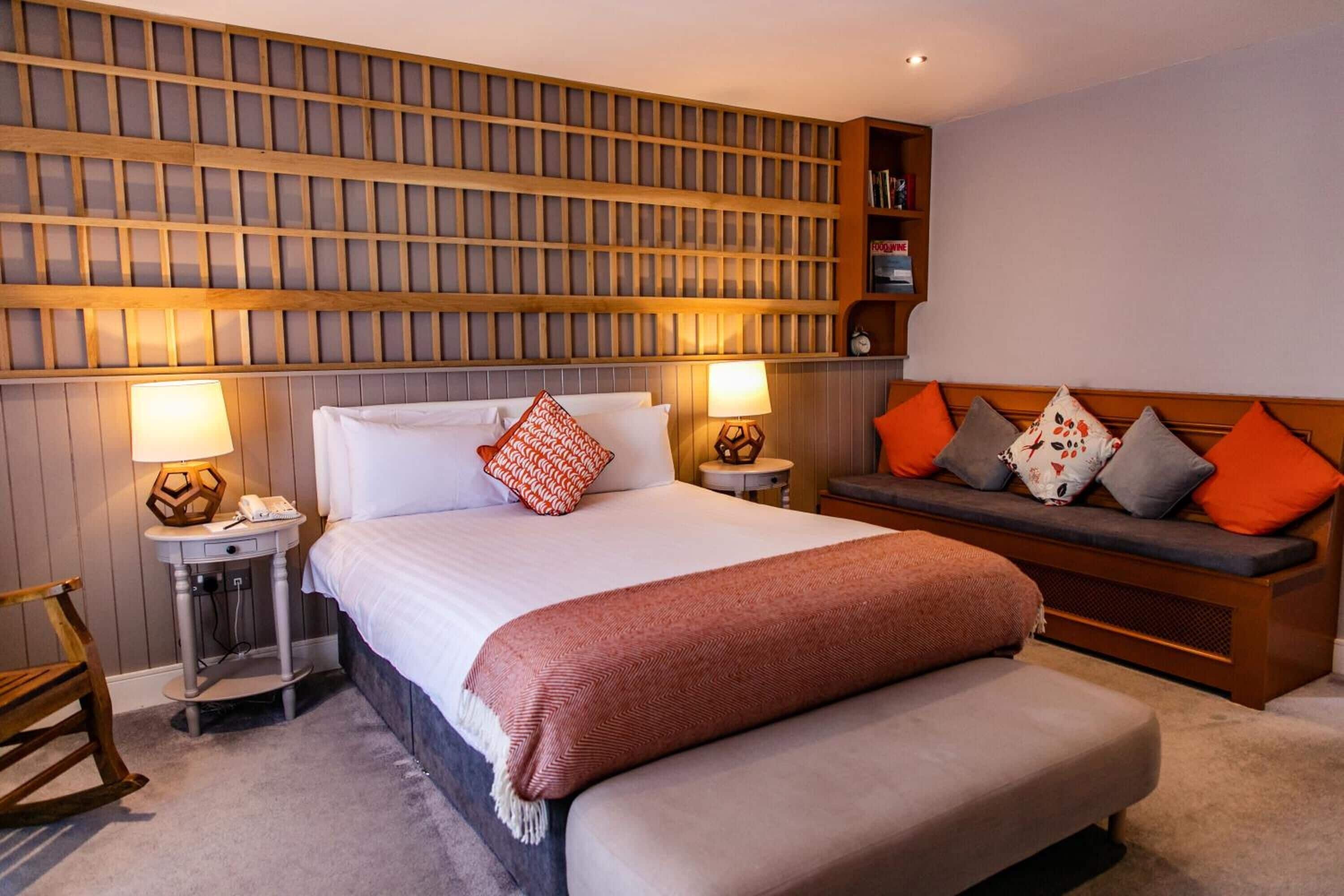 A neatly arranged hotel room with a large bed, bedside lamps, a wooden headboard, a bench with cushions, and a bookshelf.