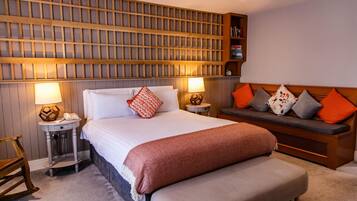 A neatly arranged hotel room with a large bed, bedside lamps, a wooden headboard, a bench with cushions, and a bookshelf.