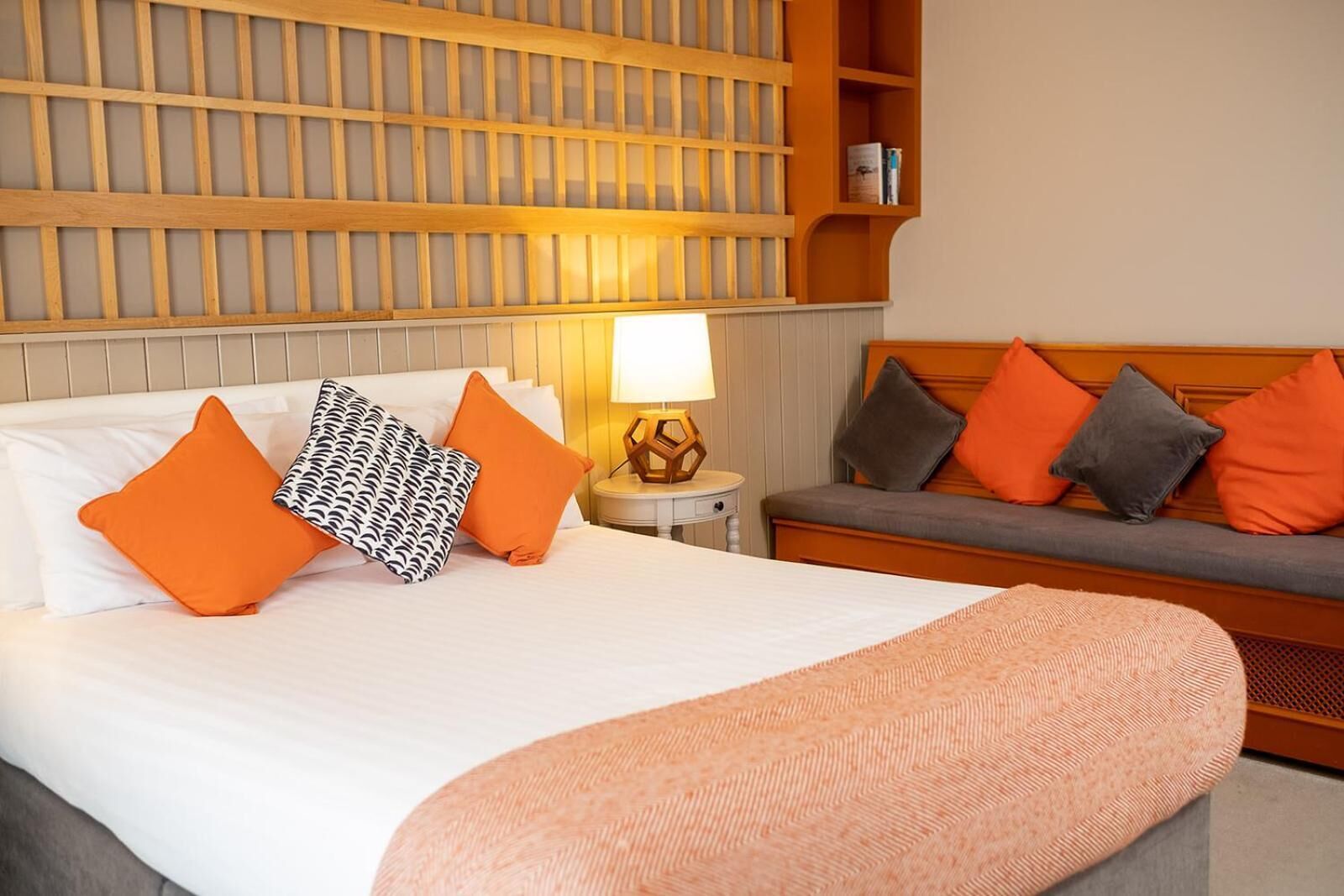 A hotel room with a bed, a nightstand with a lamp, a sofa, and a wall with a wooden panel design.