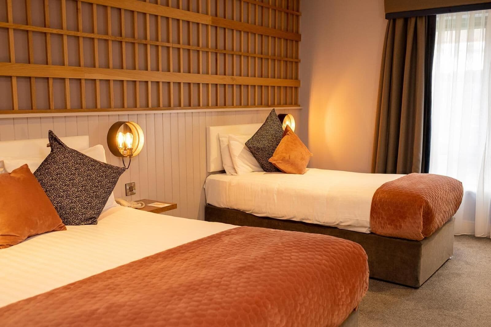 A hotel room with two beds, a wooden headboard, a nightstand with a lamp, and a window with curtains.
