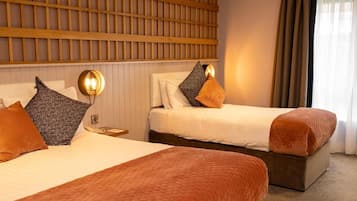 A hotel room with two beds, a wooden headboard, a nightstand with a lamp, and a window with curtains.