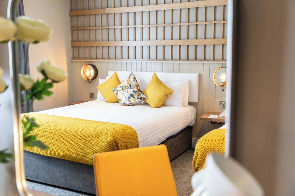 A hotel room with a bed, a nightstand, a mirror, and a wall-mounted light fixture.