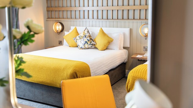 A hotel room with a bed, a nightstand, a mirror, and a wall-mounted light fixture.