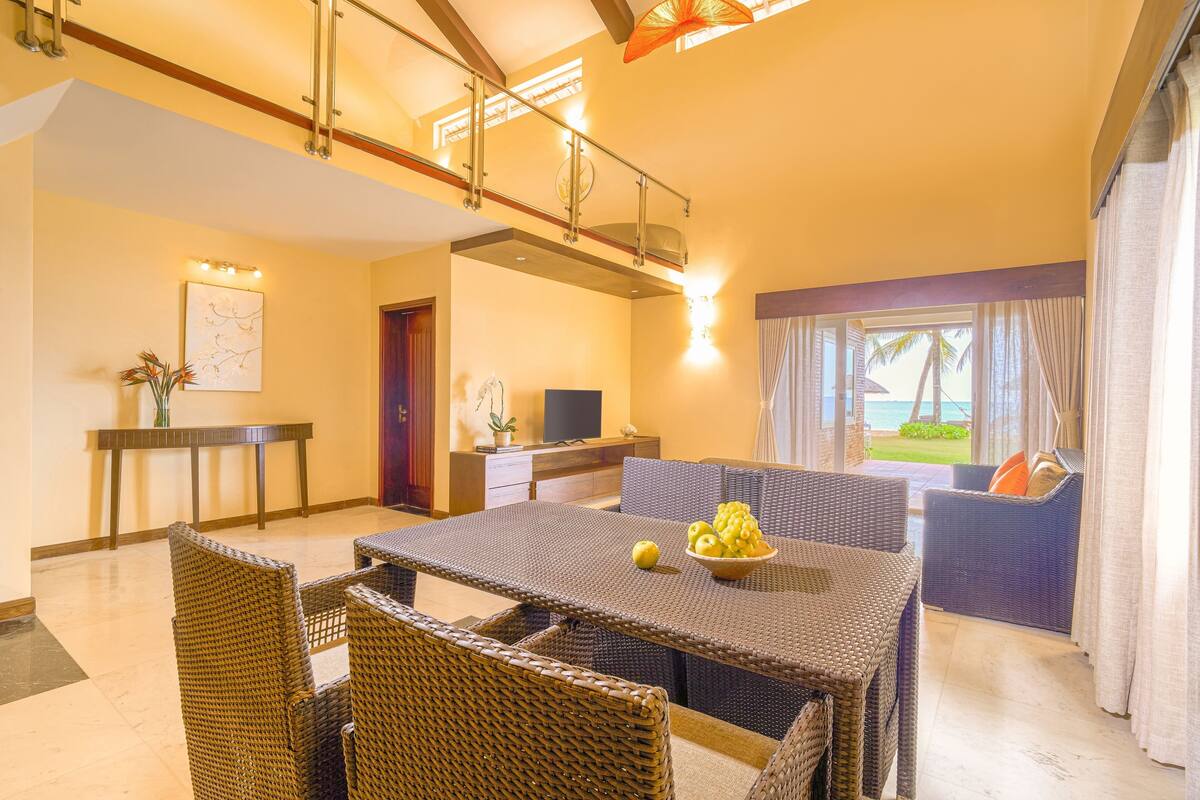 A dining area with wicker furniture, a table with a bowl of fruit, and a view of the outdoors through large windows.