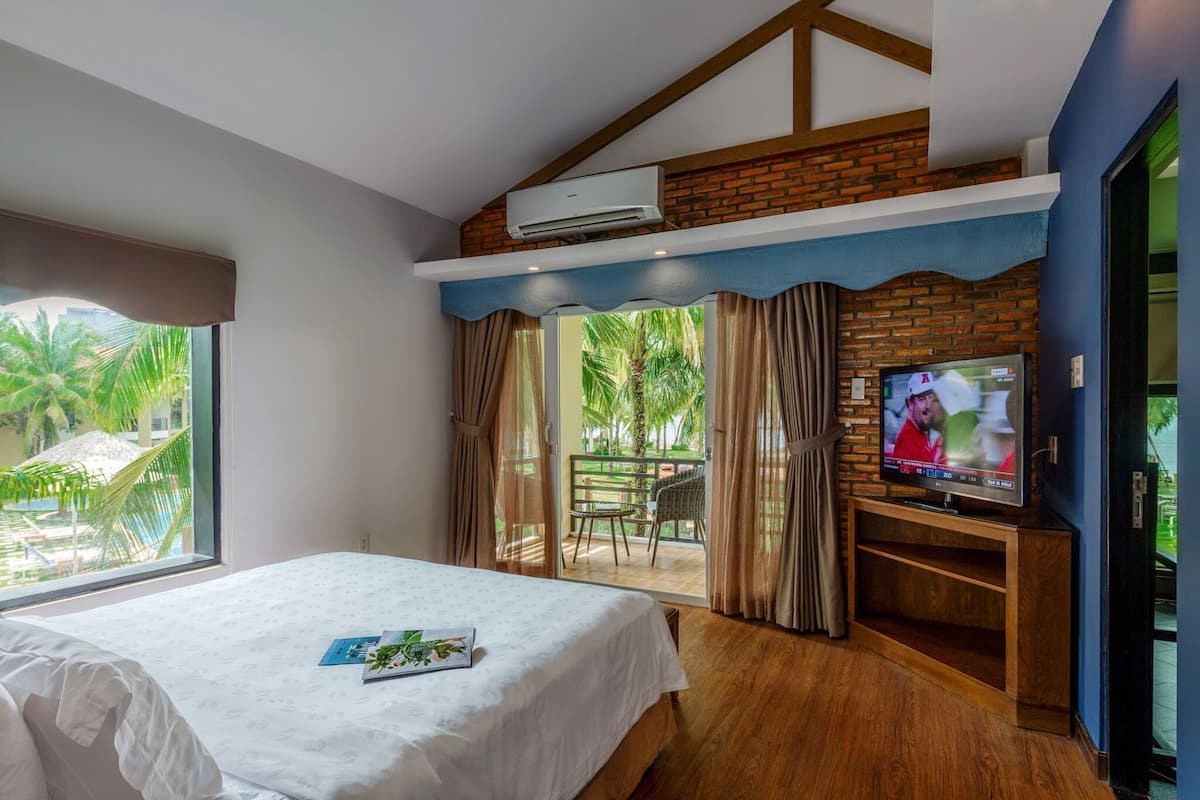 A hotel room with a bed, a TV, a balcony with a table and chairs, and a view of palm trees.