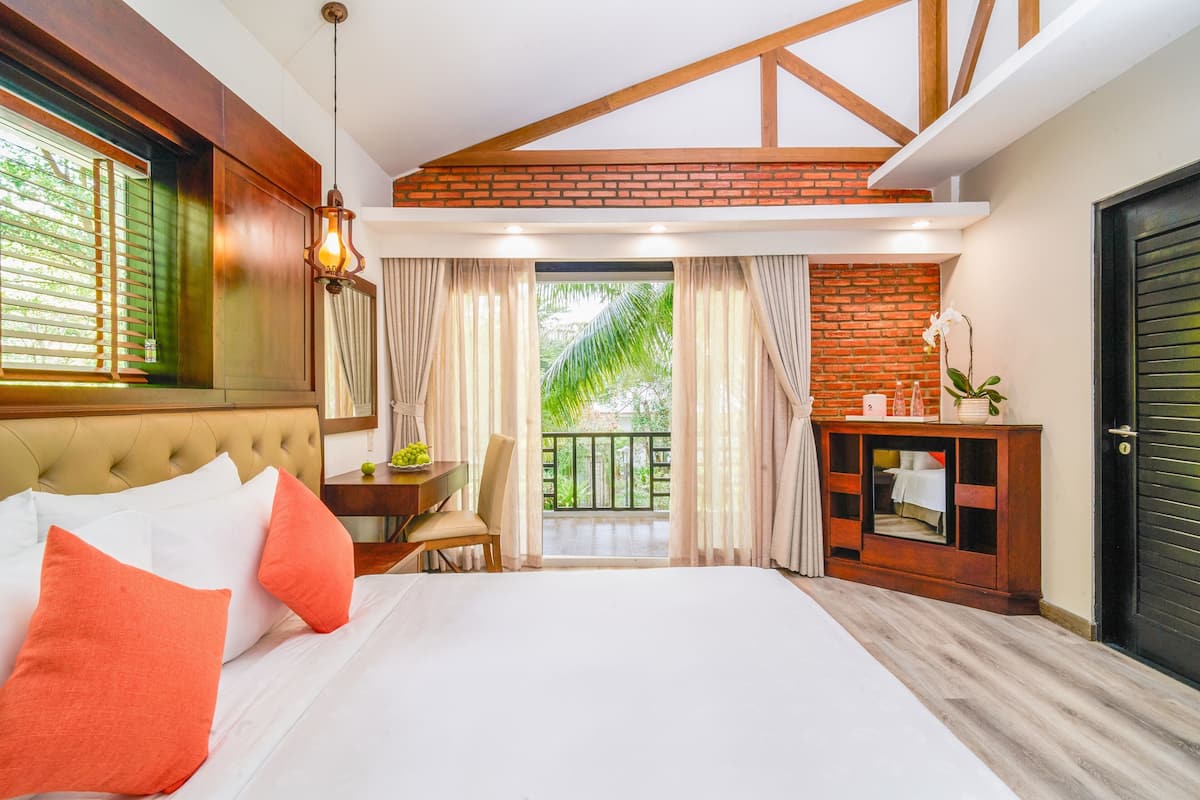 A hotel room with a large bed, a desk, a chair, a small table, a cabinet, and a balcony with a view of greenery.