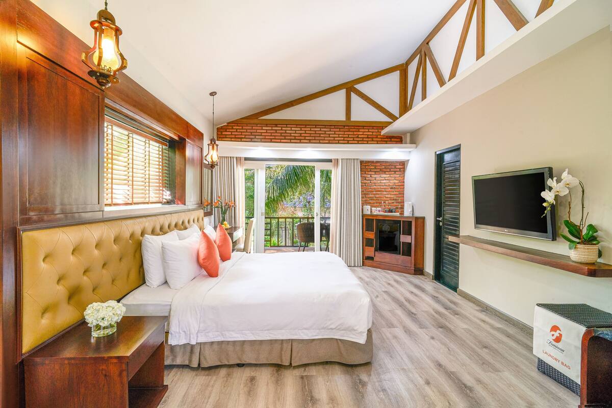 A bedroom with a large bed, wooden headboard, a TV, and a balcony with a view of greenery.