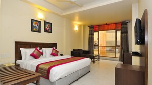 A hotel room with a large bed, a desk, a chair, a TV, and a balcony with curtains. - Shanti Villa (New Delhi)