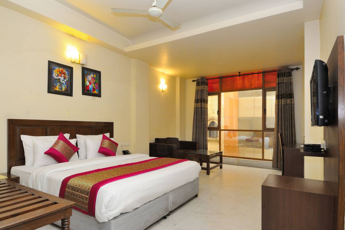 A hotel room with a large bed, a desk, a chair, a TV, and a balcony with curtains.