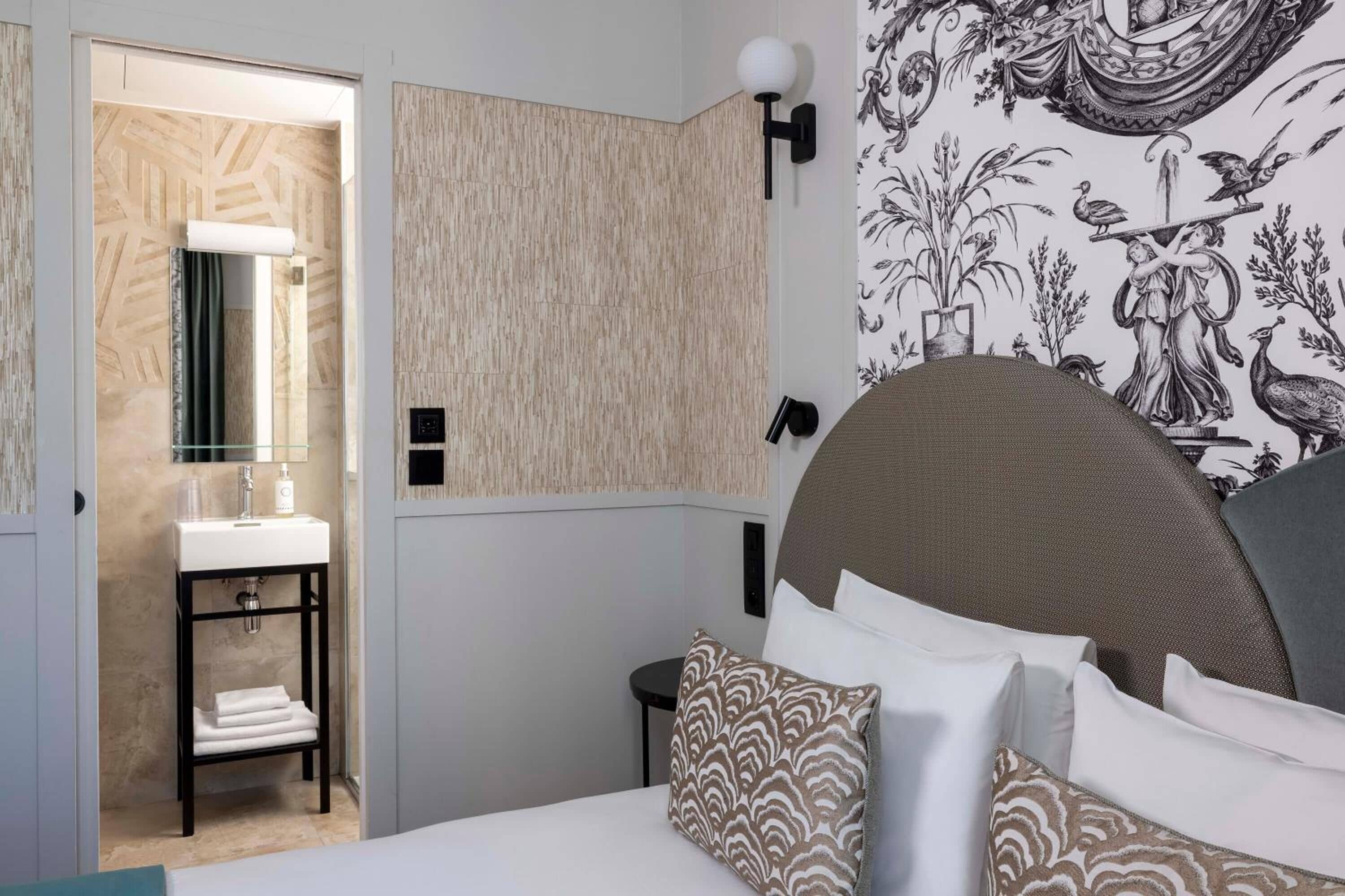 A modern hotel room with a bed, a bedside table, a wall-mounted lamp, and a bathroom visible through an open door.