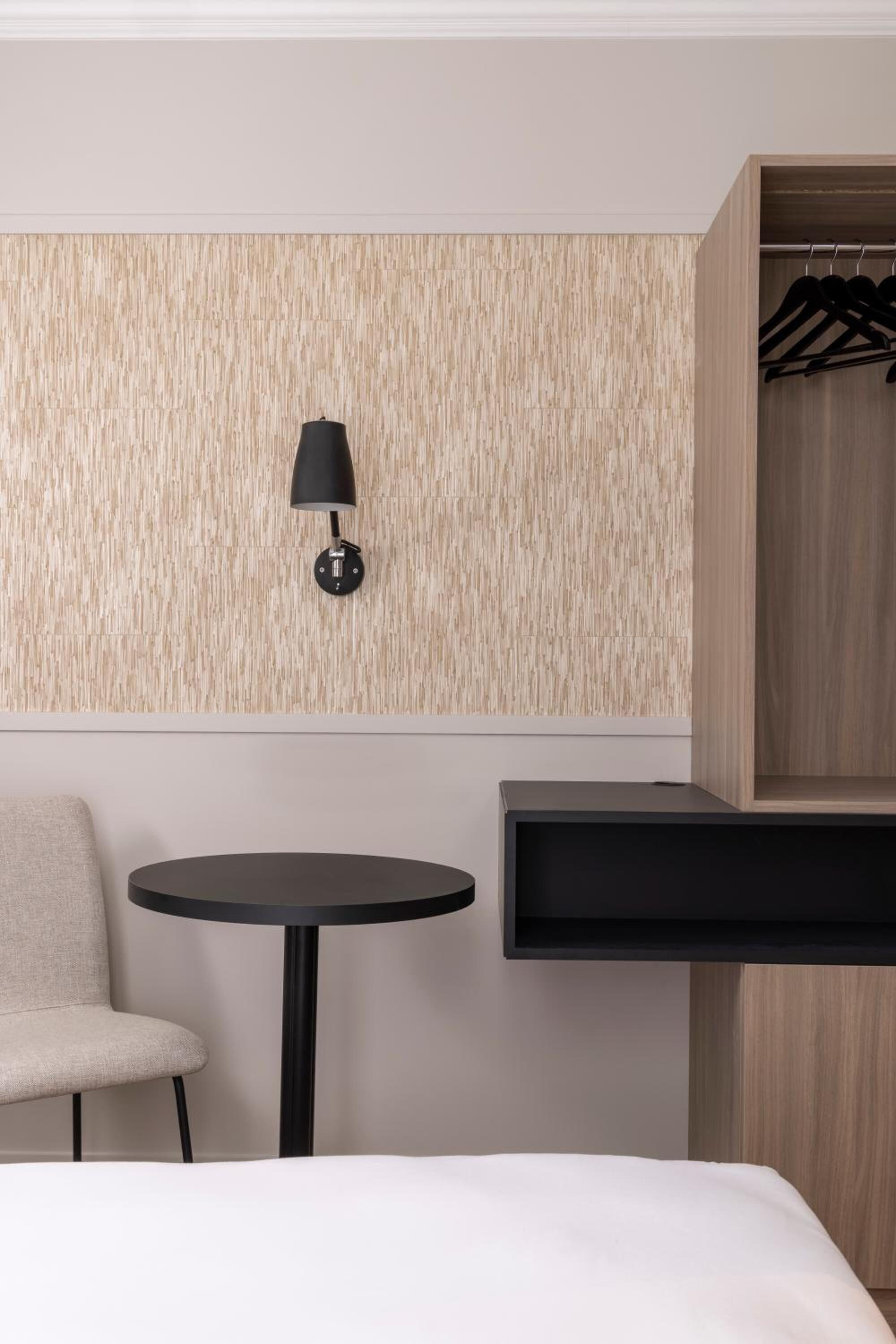 A modern hotel room with a bed, a round table, a chair, a wall-mounted lamp, and a wooden wardrobe.