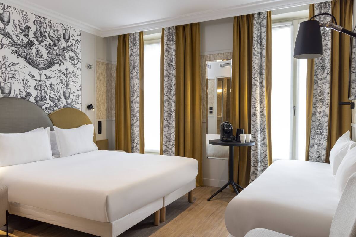 A hotel room with two beds, a small table with a lamp, and a wall with a black and white floral wallpaper.