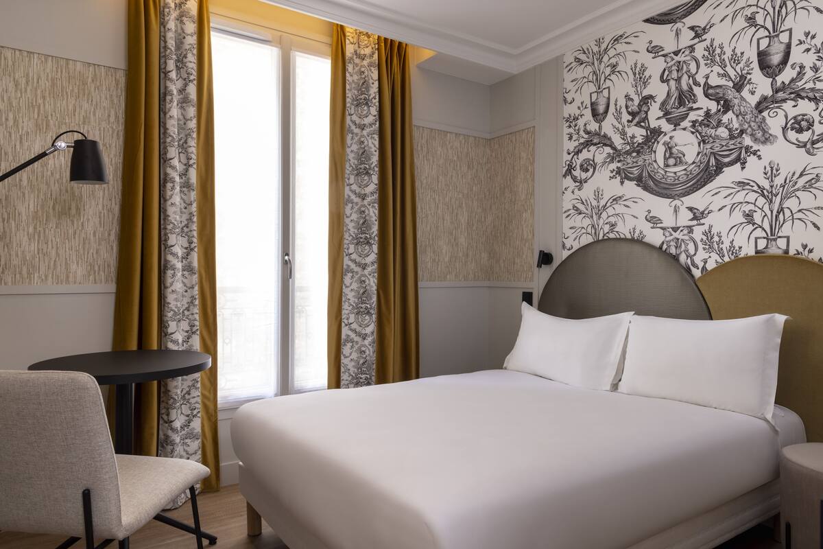 A hotel room with a bed, a chair, a small table, and a wall with a decorative pattern.