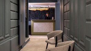 A reception area with a counter, a person behind it, and a chair in the foreground. - Grand Hotel Leveque (Paris)