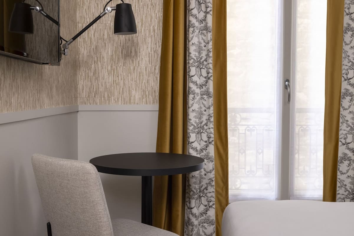 A hotel room with a bed, a round table, a chair, a lamp, and a window with curtains.