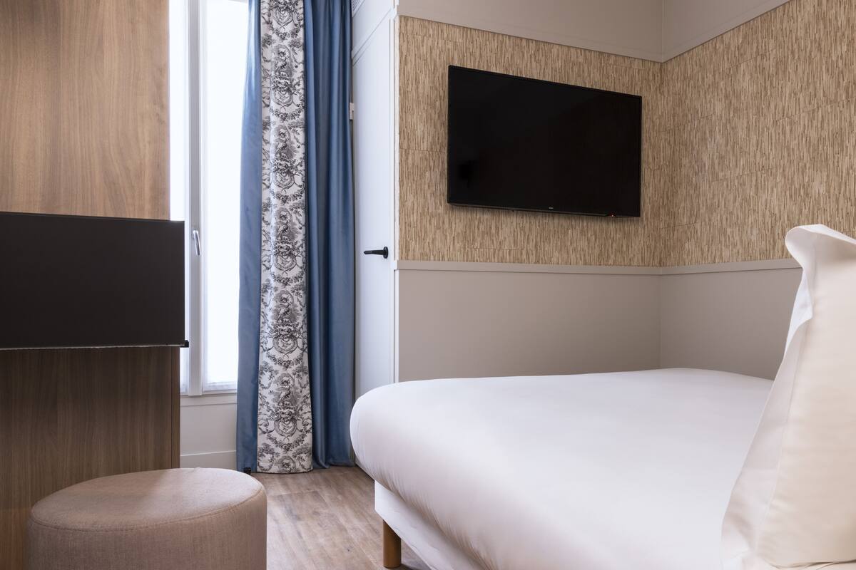 A modern hotel room with a bed, a flat-screen TV mounted on the wall, a wooden wardrobe, and a small ottoman.