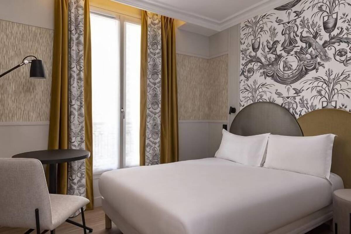 A hotel room with a bed, a chair, a small table, and a window with curtains.