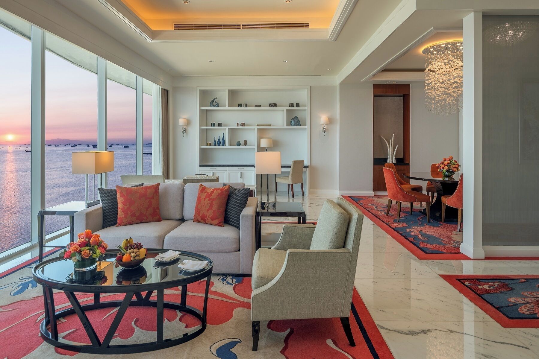 A modern living room with a large window overlooking a body of water, a round coffee table with fruit, a sofa with decorative pillows, and a chandelier.