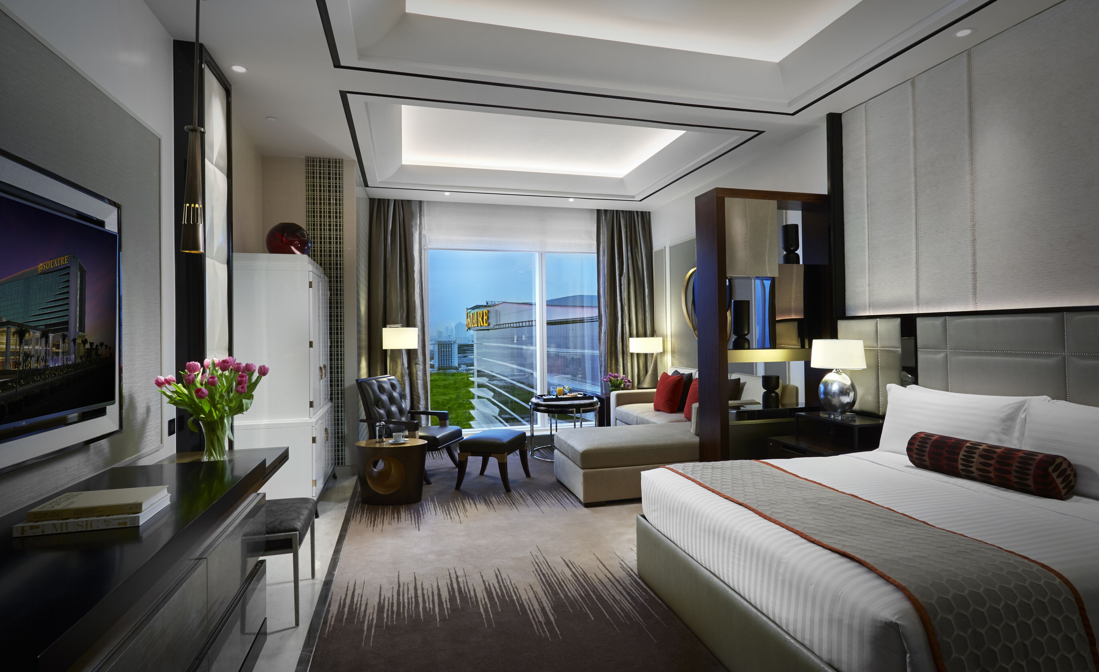 A modern hotel room with a large bed, a desk with a flat-screen TV, a seating area with a chair and a small table, and a view of a cityscape through large windows.
