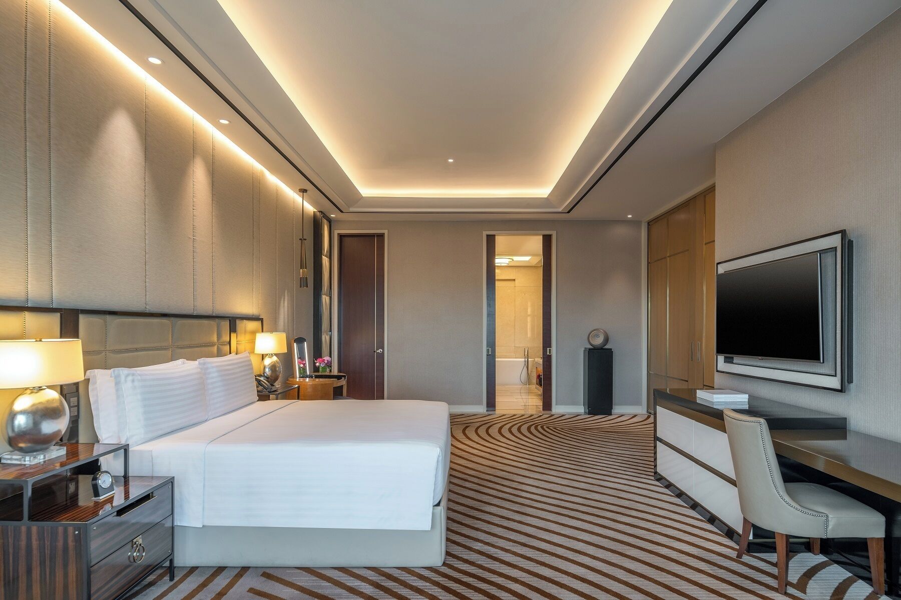 A modern hotel room with a large bed, a TV, a desk, and a bathroom visible through an open door.