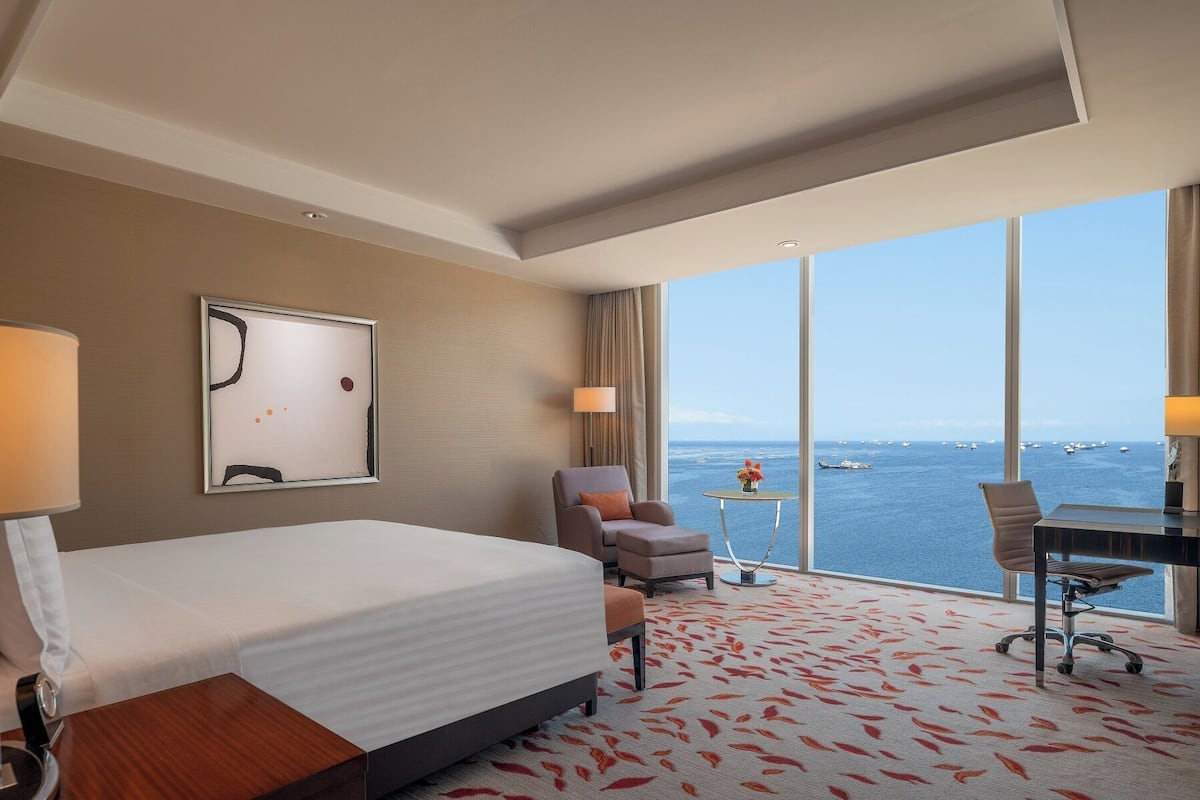 Une chambre d’hôtel comprenant un lit, deux fauteuils, un bureau et une grande fenêtre donnant sur la mer.