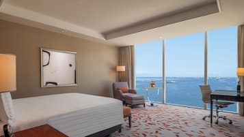 A hotel room with a bed, two armchairs, a desk, and a large window offering a view of the sea.