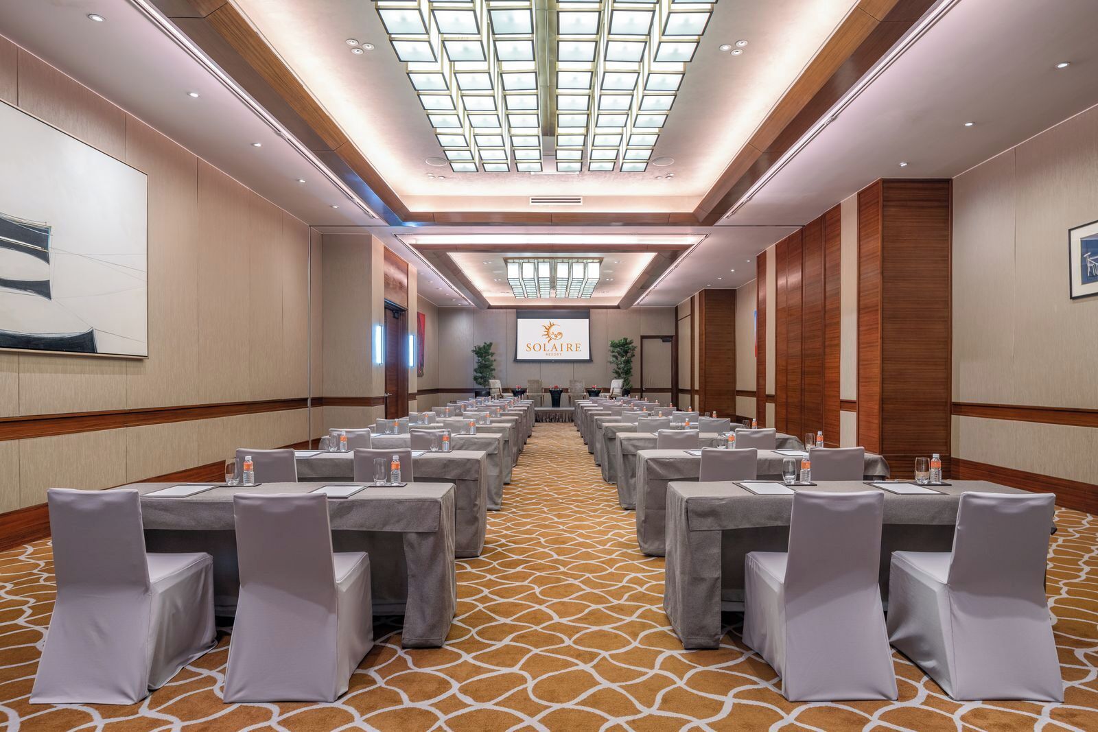 A conference room with rows of tables and chairs, a large screen at the front, and a patterned carpet.