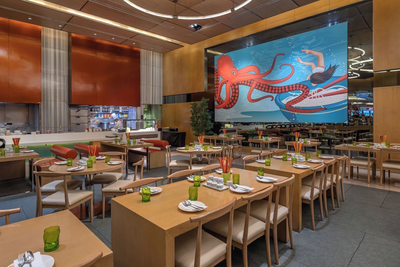 A restaurant with multiple dining tables and chairs, a large mural of an octopus on the wall, and a bar area in the background.