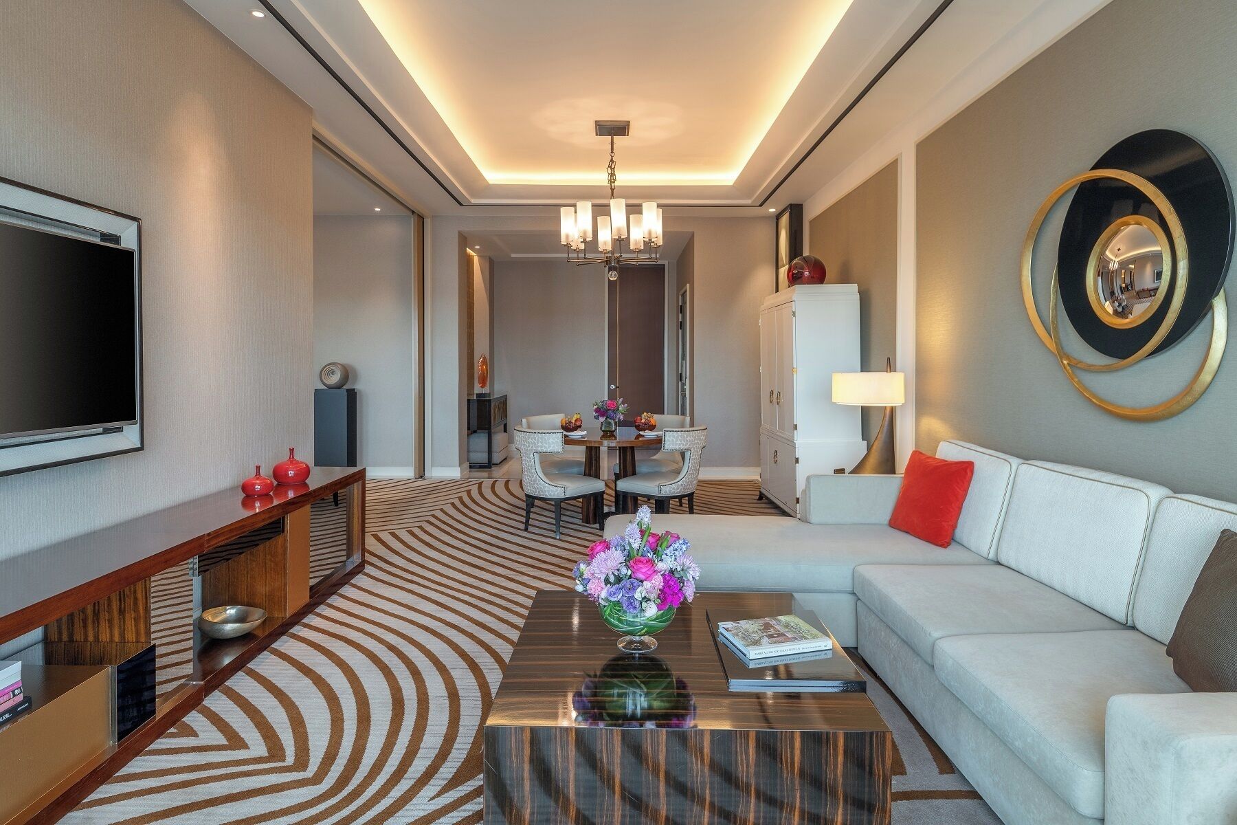 Una camera d'albergo moderna con TV a schermo piatto, una poltrona, un tavolino da caffè con un vaso di fiori, un'area pranzo illuminata da un lampadario e un grande specchio a parete.