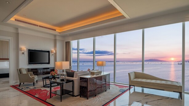 A modern hotel room with a large window offering a view of the sea at sunset, a flat-screen TV, a dining area, and a sofa.