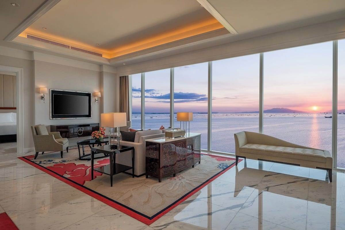 Une chambre d’hôtel moderne dotée d’une grande fenêtre offrant une vue sur la mer au coucher du soleil, d’un téléviseur à écran plat, d’un coin repas et d’un canapé.