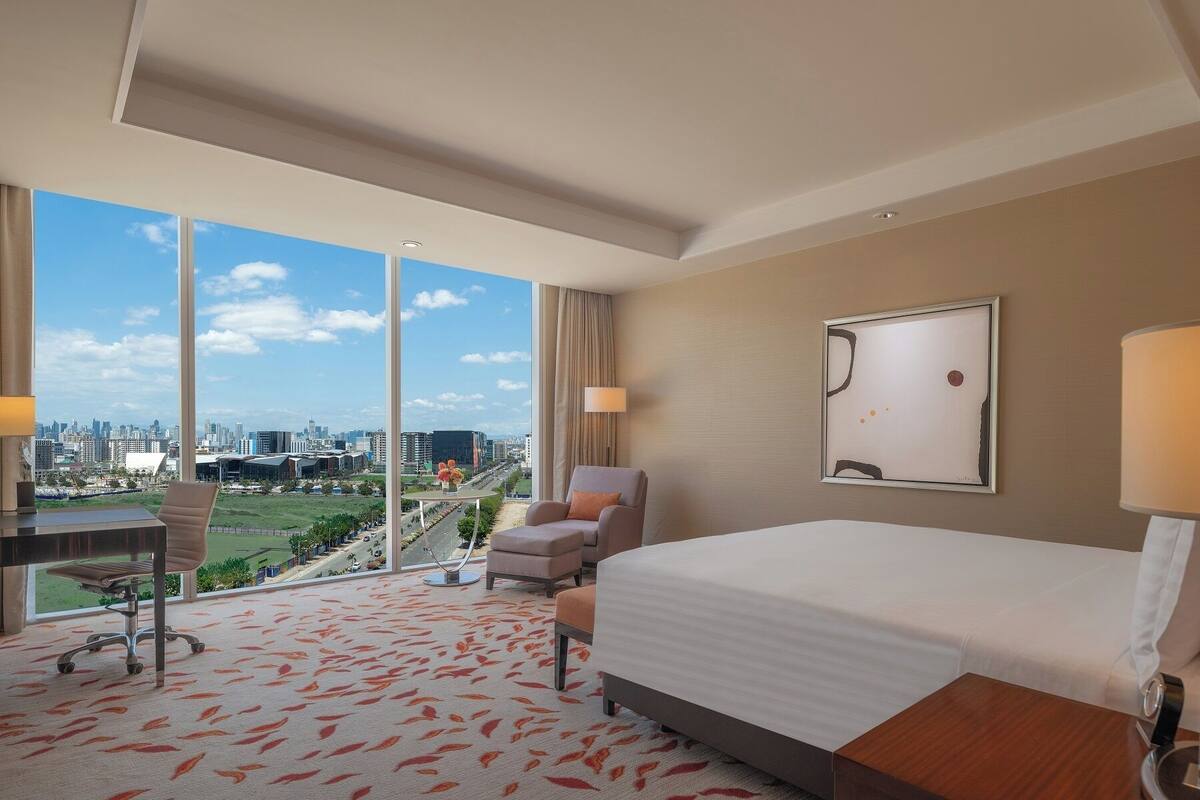 Une chambre d’hôtel avec un grand lit, un bureau et une chaise. De grandes fenêtres offrent une vue sur le paysage urbain.