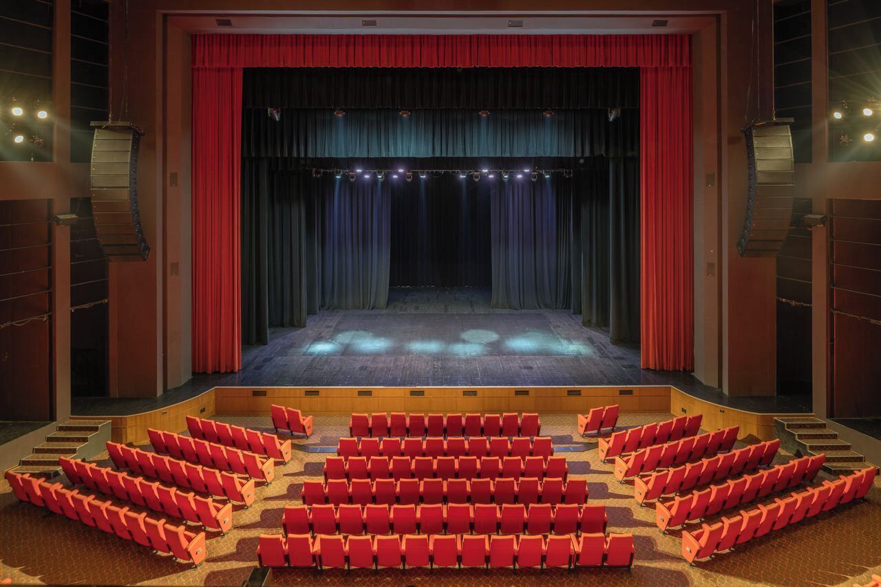 Un teatro con un escenario, cortinas rojas y hileras de butacas rojas.