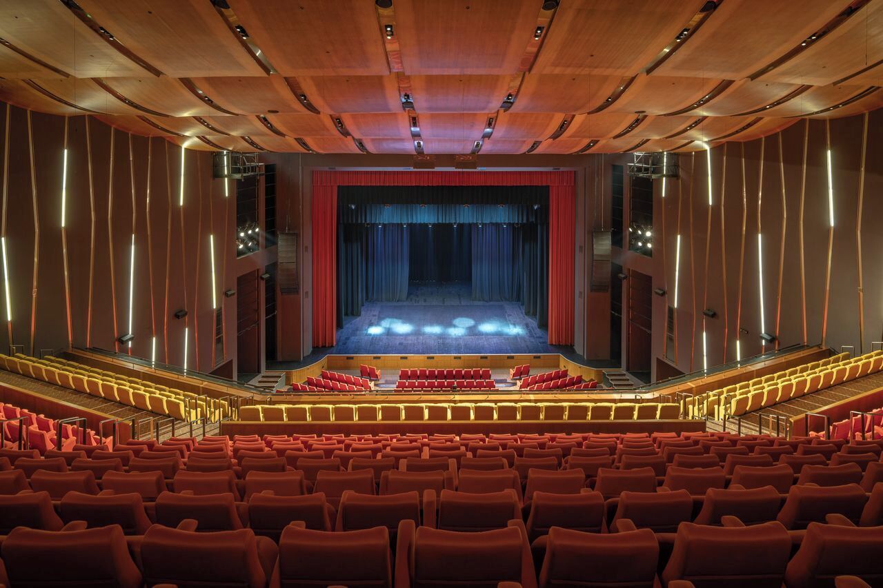 Une salle de spectacle dotée de gradins, d’une scène avec des rideaux et d’un plafond en bois agrémenté de luminaires encastrés.