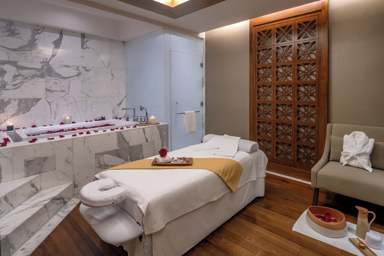 A spa room with a massage bed, a bathtub with rose petals, a wooden partition, and a seating area with towels.