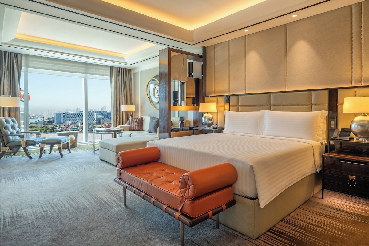 Une chambre d’hôtel moderne dotée d’un grand lit, d’un coin salon avec un canapé et des fauteuils, offrant une vue sur la ville, et dont les fenêtres donnent sur le panorama urbain.