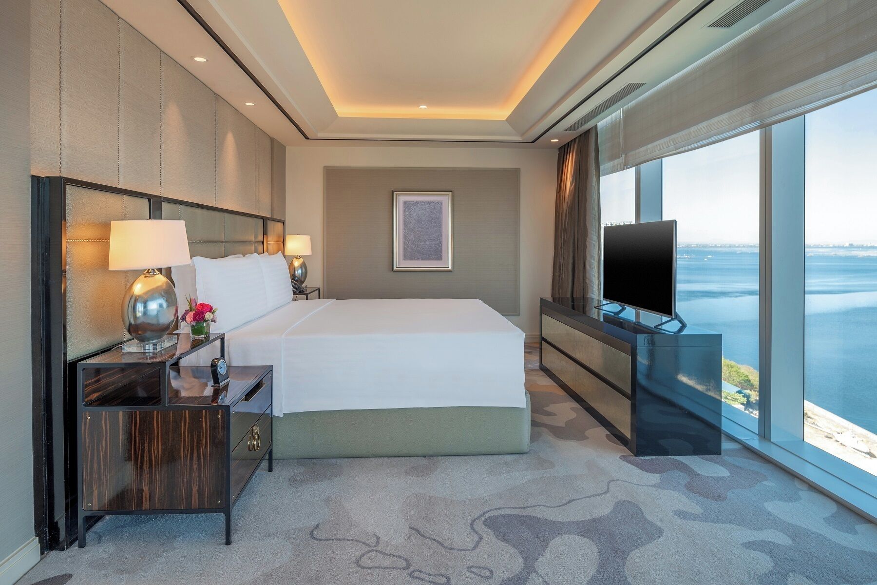 A hotel room with a large bed, a TV, a desk with a lamp, and a view of the sea.