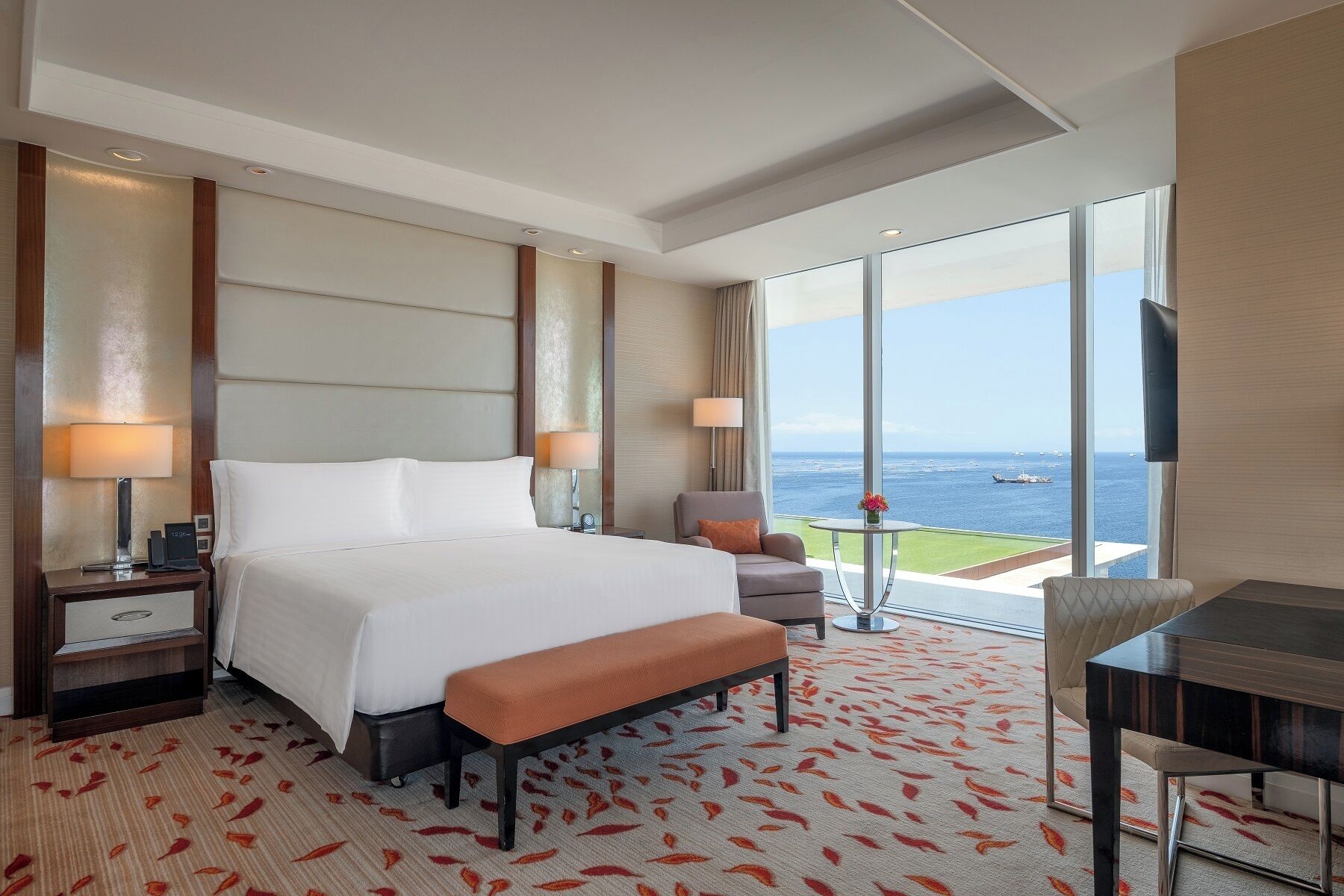 A hotel room with a large bed, a desk, a chair, and a view of the sea.