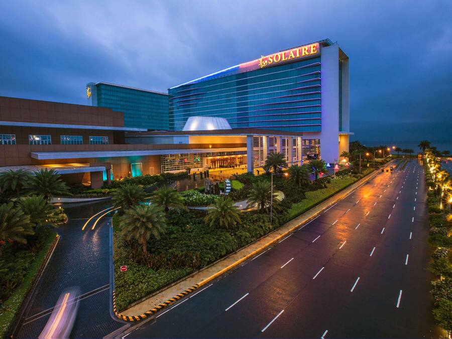 A modern hotel building with a prominent sign, a well-maintained road, and landscaped areas with palm trees.