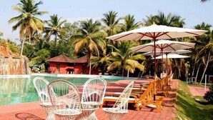 A pool area with lounge chairs and umbrellas, surrounded by palm trees and a small building. - Club Mahindra Kumarakom (Kottayam)