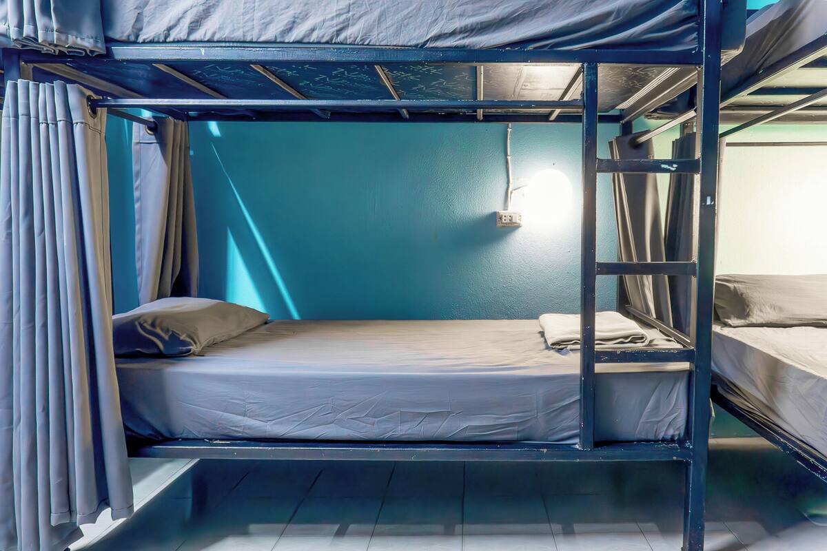 Ein Schlafraum mit Etagenbett, einem Einzelbett und einer steilen Leiter, einer Steckdose an der Wand und Vorhängen auf der linken Seite.