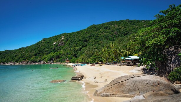 Une plage bordée d’une eau turquoise et limpide, de grands rochers et, en arrière-plan, de collines verdoyantes.