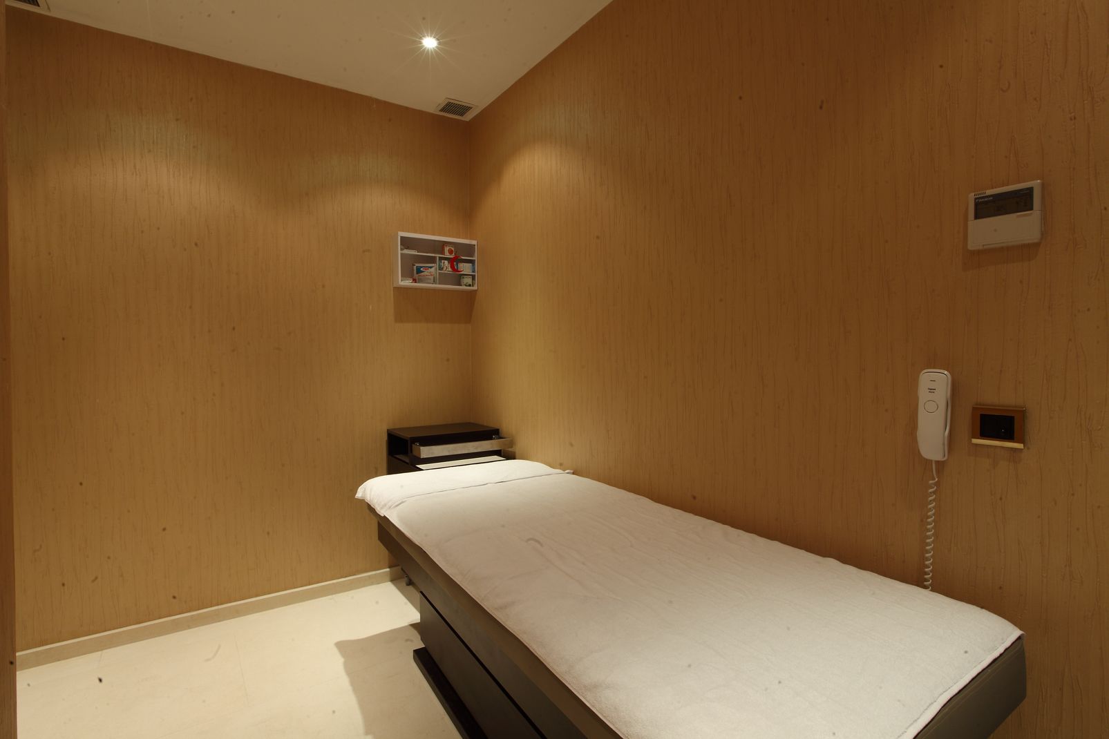 Una habitación pequeña con cama individual, paredes de madera, una mesita de noche, un estante con libros y un teléfono.