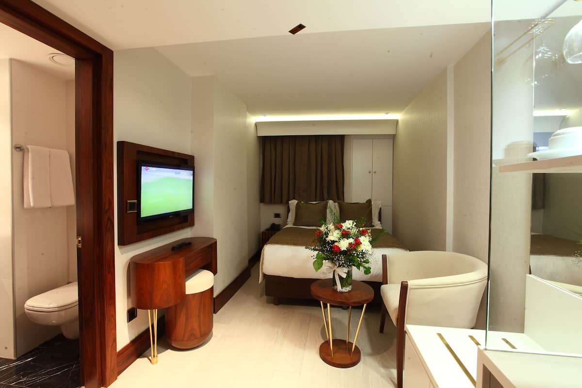 Une chambre d’hôtel comprenant un lit, une télévision, un bureau, une chaise, une petite table avec un vase de fleurs et une vitrine.