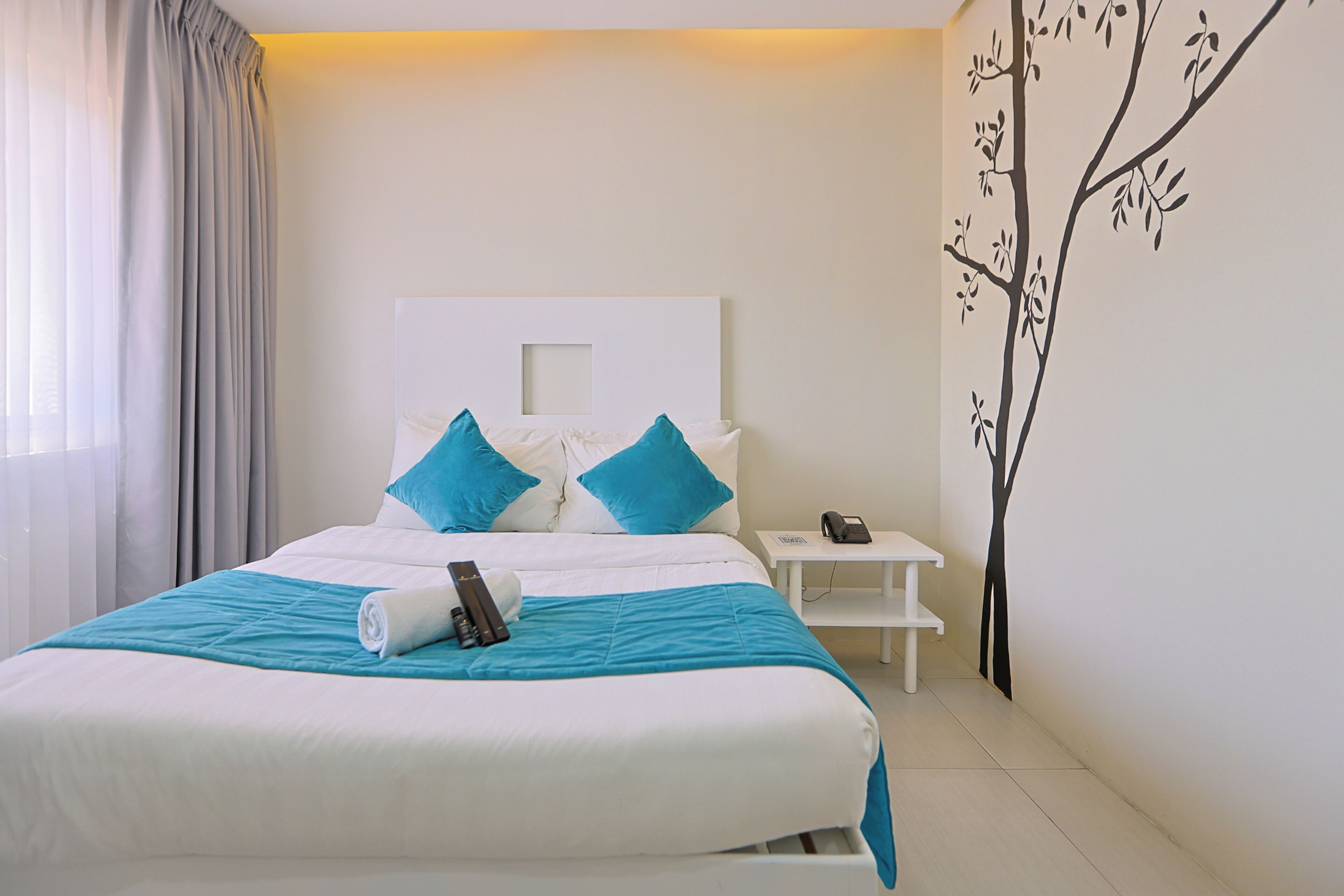 A neatly made bed with a blue and white bedspread, a white nightstand with a phone, and a wall with a tree mural.