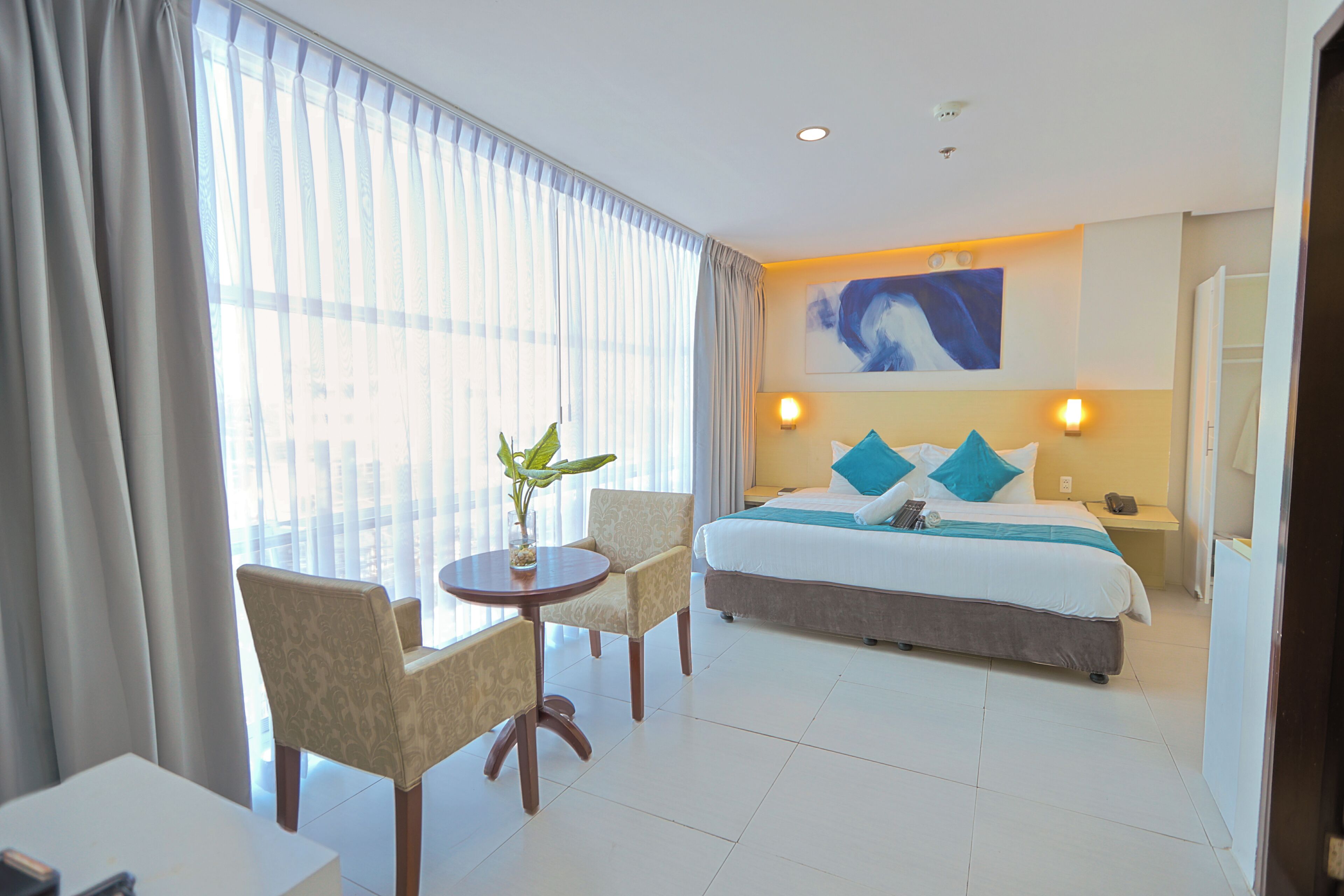 A hotel room with a bed, two chairs, a small table with a plant, and a painting on the wall.