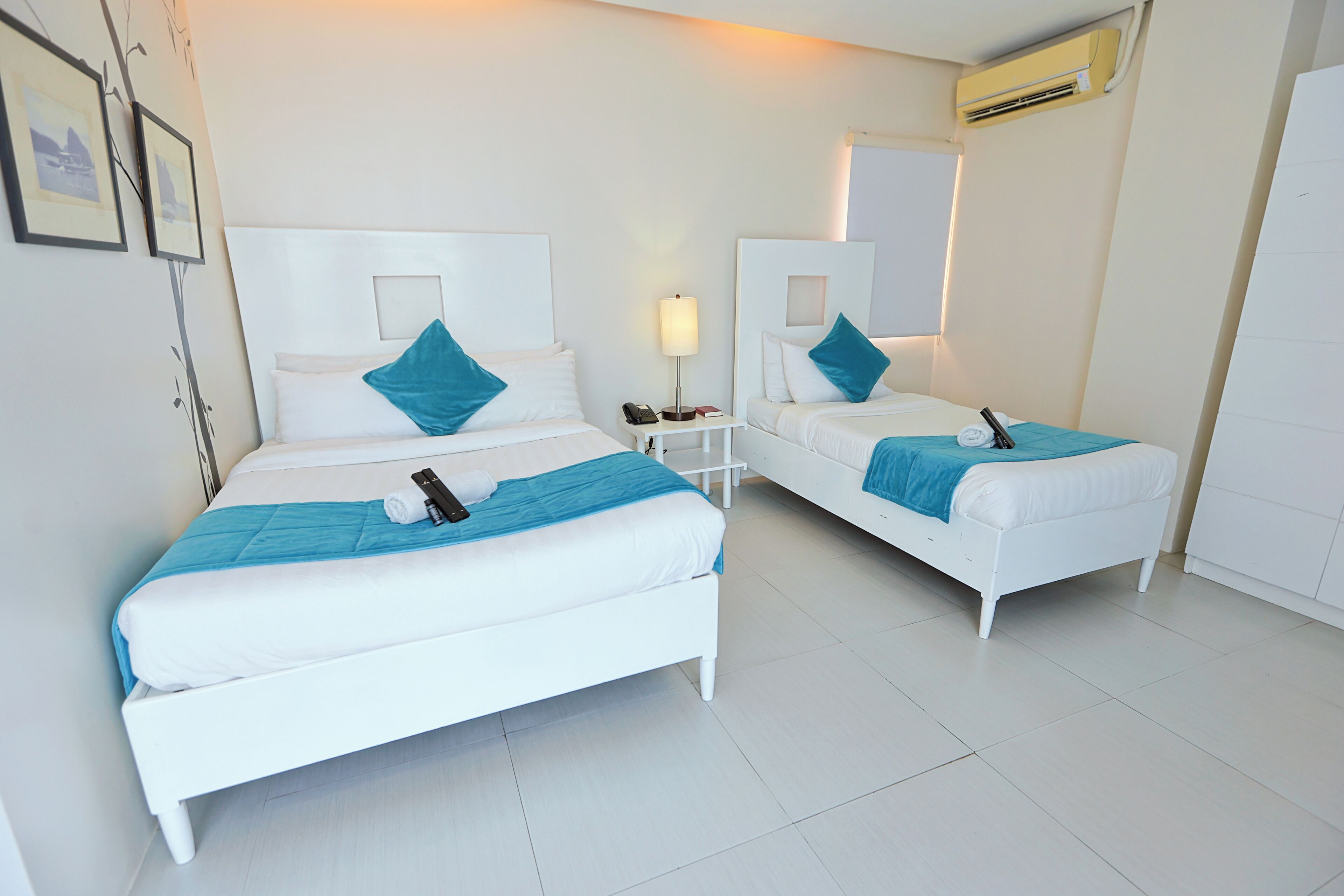 A hotel room with two white beds, each with a blue pillow and blanket, a bedside table with a lamp, and an air conditioning unit on the wall.