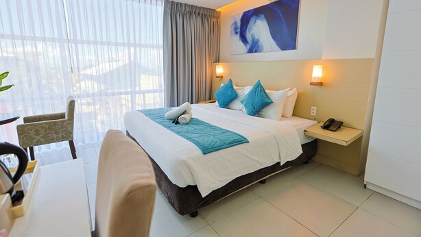A hotel room with a bed, a desk, a chair, a nightstand with a phone, and a painting on the wall. - Holiday Suites Business District (Empire Suites Hotel) (Puerto Princesa)